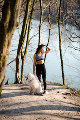 Full height portrait of a young sports girl with the dog on a sunny spring morning in the park. A woman stands next to her dog on a hill near the lake