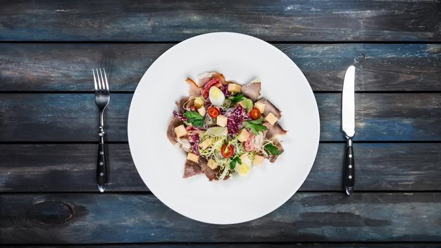Rotating salad on a plate. Mixed lettuce, tomatoes, turkey smoked, smoked duck, dried horsemeat. Served with cutlery on a colored wooden background. Top view