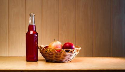 Glass bottle with red apple cider and apples in a basket on a wooden background with copy space.