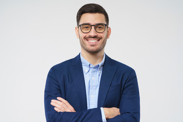 Close-up portrait of confident young businessman wearing blue blazer and smart casual shirt, smiling at camera, holding arms crossed, standing isolated on gray background