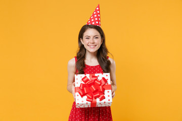 Smiling young brunette woman girl in red summer dress, birthday hat posing isolated on yellow background. Birthday holiday concept. Mock up copy space. Hold white red present box with gift ribbon bow.