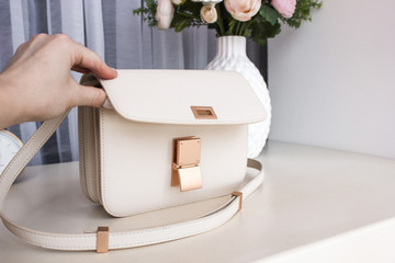 White women's leather bag with on a white table with a vase, flowers and a clock in the background