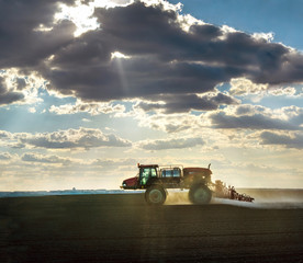 a self-propelled sprayer in the field cultivates arable land against the fantastic light from under the clouds - obrazy, fototapety, plakaty