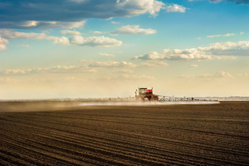 Big red sprayer in the arable field makes fertilizers in early spring , backlit jet, rear view with evening clouds sky