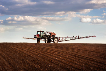Big red sprayer with long arms at the arable field makes fertilizers in early spring view with clouds - obrazy, fototapety, plakaty