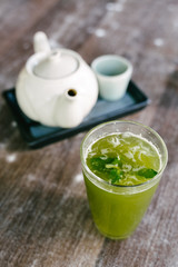 Refreshing glass of homemade lemonade decorated with mint leaves and a ceramic tea set with teapot and small cup on a ceramic tray as a background, placed on a wooden table