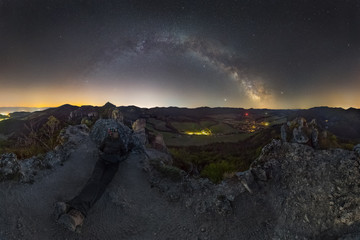 Panorama night landscape with a picturesque village in the valley between the mountains. The Milky Wax rises above the horizon in the night sky. A man sits on top with a respirator and looks at sky.