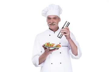 Old male cook holding a plate with vegetables salad and pepper on white background
