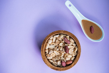Peanuts in a wooden bowl and penunt butter in a spoon on purple background. Healthy snack. Top view.
