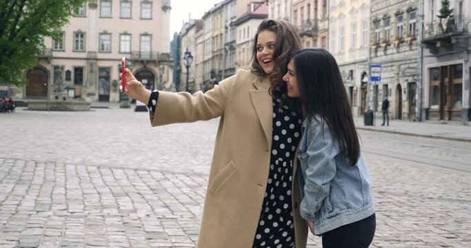 Girls taking selfie self portrait photos on smartphone. Models posing on street background. Female showing positive face emotions