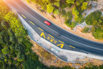 Aerial view of road in beautiful green forest at sunset in summer. Colorful landscape with red car on the roadway, trees in spring. Top view from drone of highway in Croatia. View from above. Travel