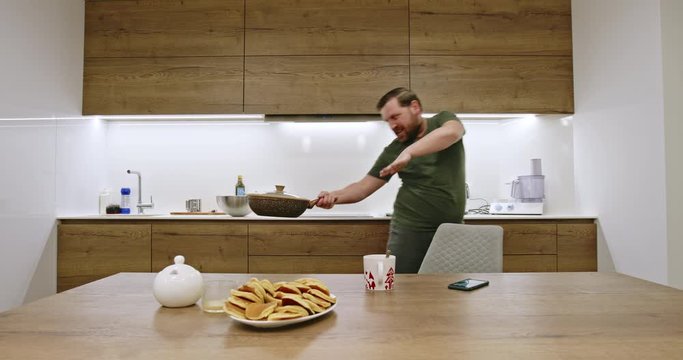 Happy man cooking dinner and dancing before smelling the food in the frying pan and starting to snack from a bowl alongside viewed across a table with cookies