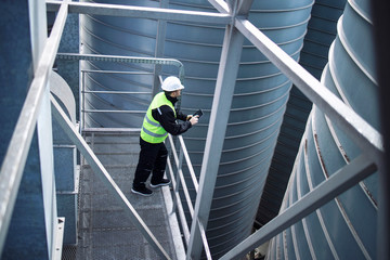 Factory silos worker standing on metal platform between industrial storage tanks and looking at...