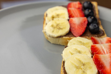 Sandwiches with fruits on a grey plate. Concept of delicious and healthy breakfast. Toasts with chocolate topping, banana, strawberry and blueberry.