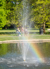 Three girls (out of focus) sitting on a bench at Pinner Memorial Park, Pinner, Middlesex UK. Photographed through the water fountain in the duck pond, with the reflection of the sun creating a rainbow