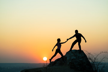 Man and woman hikers helping each other to climb a big stone at sunset in mountains. Couple...