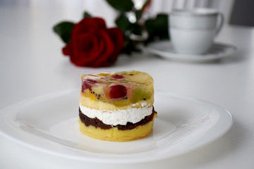 Layer cake with fruits and cream on a white plate in front of red rose and service of cup and plate
