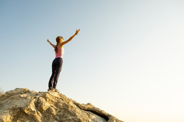 Young woman hiker standing alone on big stone in mountains. Female tourist raising her hands up on high rock in morning nature. Tourism, traveling and healthy lifestyle concept.