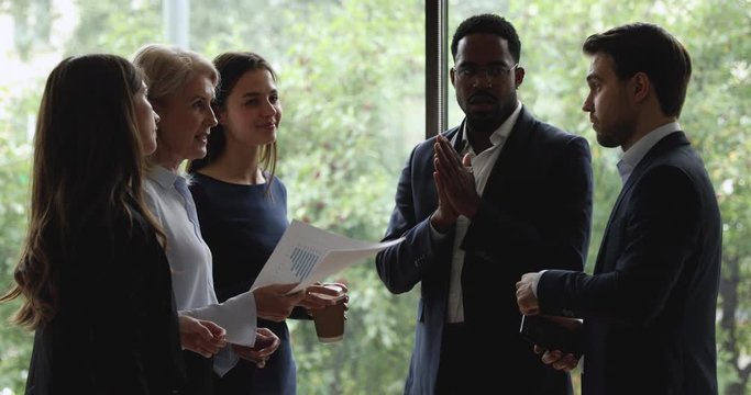Different ages successful diverse business people standing at big window, holding casual conversation at working space. Multiracial younger and older professional workers discussing working issues.
