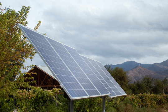 Dos paneles fotovoltaicos de uso dom&eacute;stico en medio de un cultivo de parras en una granja org&aacute;nica. Es un d&iacute;a nublado. Detr&aacute;s se ve el techo de una bodega y en el fondo, las monta&ntilde;as.