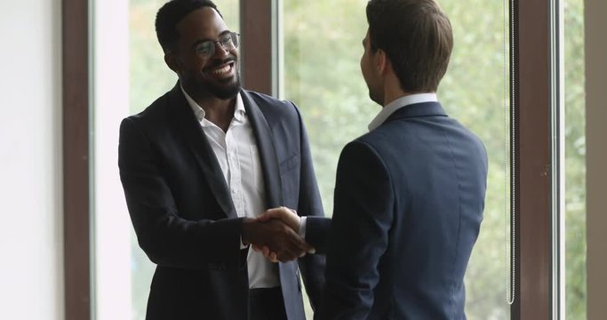 Happy biracial manager in eyewear shaking hands with motivated partner near huge window in modern working space. Smiling young multiethnic businessmen making agreement, establishing partnership.