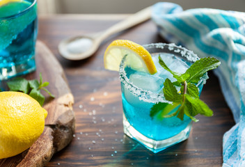 Alcoholic cocktail on a wooden table surrounded by ingredients such as lemon, salt, sugar, ice