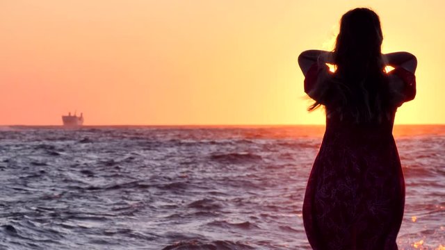 Silhouette of slender young girl at sunset, dawn, against background of sun, sky and sea. A girl in dress with long hair slowly raises her hands to sun, says goodbye to sun, touches, plays with hair.