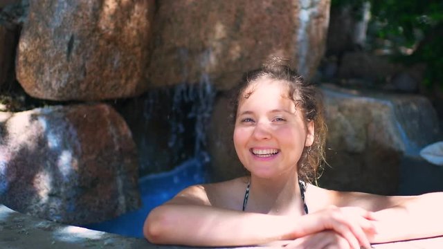 Young happy smiling woman inside Japanese spa stone pool waterfall plunge with colorful blue hot springs mineral water in Japan onsen in slow motion