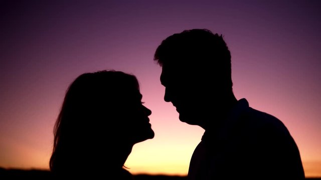 Lovely couple silhouettes kissing at the sunset. Man and woman are laughing and kiss each other's noses. The sky is violet and yellow because of the sunset.