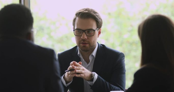 Successful young businessman in eyewear explaining business growth strategy to interested multiracial colleagues in office. Skilled male manager wearing suit, holding negotiations with coworkers.