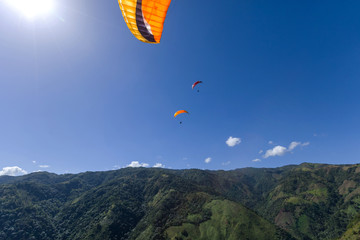 Paraglider flying in blue sky above green forests mountains landscape with sun shining