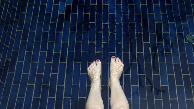 woman swings bare legs with red manicure sitting in transparent swimming pool water against blue tile bottom close upper view
