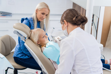Obraz premium The dentist cleaning the girl's teeth with a special tool, the girl is looking at the doctor with her beautiful blue eyes