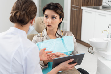 Fototapeta premium A young worried woman sitting in a dental chair and looking at her doctor, while the dentist is explaining something to her using modern technology