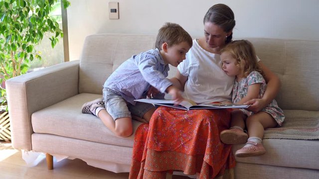 Young mother reading a book to her two children and having fun at home during quarantine