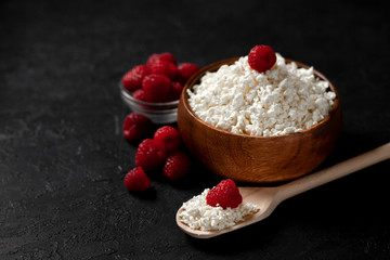 fresh homemade cottage cheese in a wooden bowl with raspberries on a dark background, healthy food on a black table