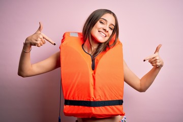 Young beautiful brunette woman wearing orange lifejacket over isolated pink background looking...