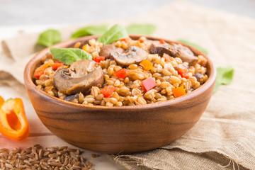 Spelt (dinkel wheat) porridge with vegetables and mushrooms in wooden bowl on a white wooden background and linen textile. Side view, selective focus.