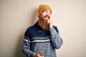 Handsome Irish redhead man with beard wearing winter sweater and wool hat with hand on chin thinking about question, pensive expression. Smiling with thoughtful face. Doubt concept.