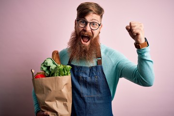Redhead Irish supermarket worker man with beard wearing apron and holding fresh groceries screaming proud and celebrating victory and success very excited, cheering emotion - obrazy, fototapety, plakaty