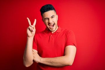 Young handsome man wearing red casual polo standing over isolated background smiling with happy face winking at the camera doing victory sign. Number two.