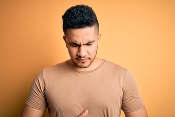 Young handsome man wearing casual t-shirt standing over isolated yellow background with hand on stomach because indigestion, painful illness feeling unwell. Ache concept.