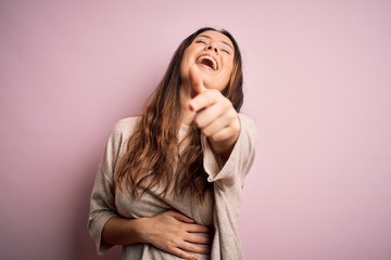Young beautiful brunette woman wearing casual sweater standing over pink background laughing at you, pointing finger to the camera with hand over body, shame expression