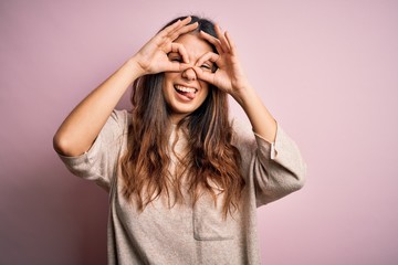 Young beautiful brunette woman wearing casual sweater standing over pink background doing ok gesture like binoculars sticking tongue out, eyes looking through fingers. Crazy expression.