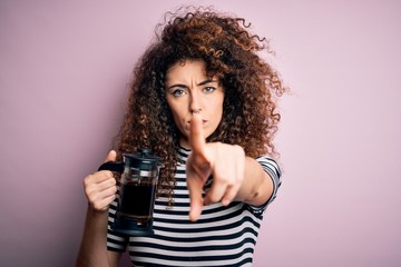 Young beautiful woman with curly hair and piercing doing coffee holding french coffeemaker pointing with finger to the camera and to you, hand sign, positive and confident gesture from the front