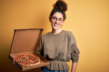 Young beautiful woman with curly hair and piercing holding delivery box with Italian pizza with a happy face standing and smiling with a confident smile showing teeth