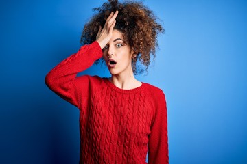 Young beautiful woman with curly hair and piercing wearing casual red sweater surprised with hand...