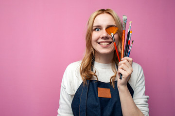 girl artist holds brushes and a palette and smiles on a pink background, student of art school, profession of an artist