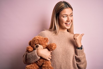 Young beautiful woman holding teddy bear standing over isolated pink background pointing and...