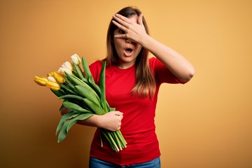 Young blonde woman holding romantic bouquet of tulips flowers over yellow background peeking in shock covering face and eyes with hand, looking through fingers with embarrassed expression.